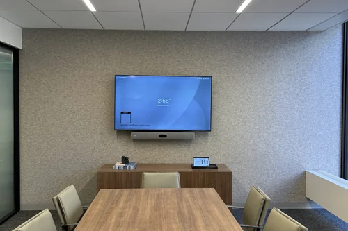 A conference room with one wall display and a video bar below it, a touch controller and a camera on the credenza, and a wood table under recessed ceiling lighting.