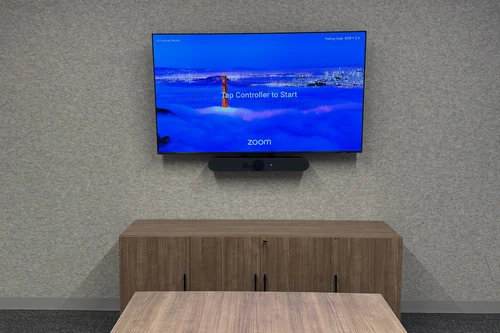 A small conference room with a wall display, a video bar beneath it, and a Logitech touch controller and remote on the table.