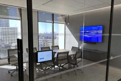 A glass-walled meeting room seen from the corridor, with a wall display, a video bar beneath it, and a touch controller on the table.
